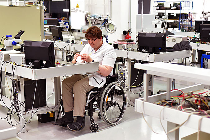 En man sitter i rullstol och arbetar vid en bänk i nån slags teknisk produktion eller repartionsverkstad.